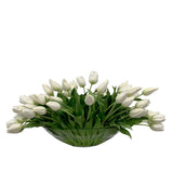 Large faux white tulip centerpiece arranged in a boat shaped clear glass vase with lush green foliage