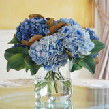 Blue hydrangea faux floral arrangement with magnolia leaves in a clear glass vase
