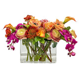Colorful faux floral arrangement with ranunculus orchids roses and calla lilies styled in a clear rectangular glass planter.