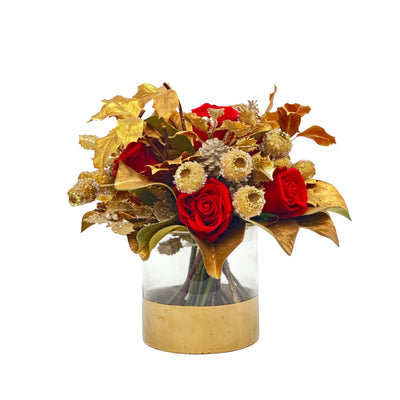 Holiday floral decor showcasing romantic faux red roses and goldleaf berry sprays, jewel pod branches, and leaves in a clear glass cylinder vase with gold base on a white background