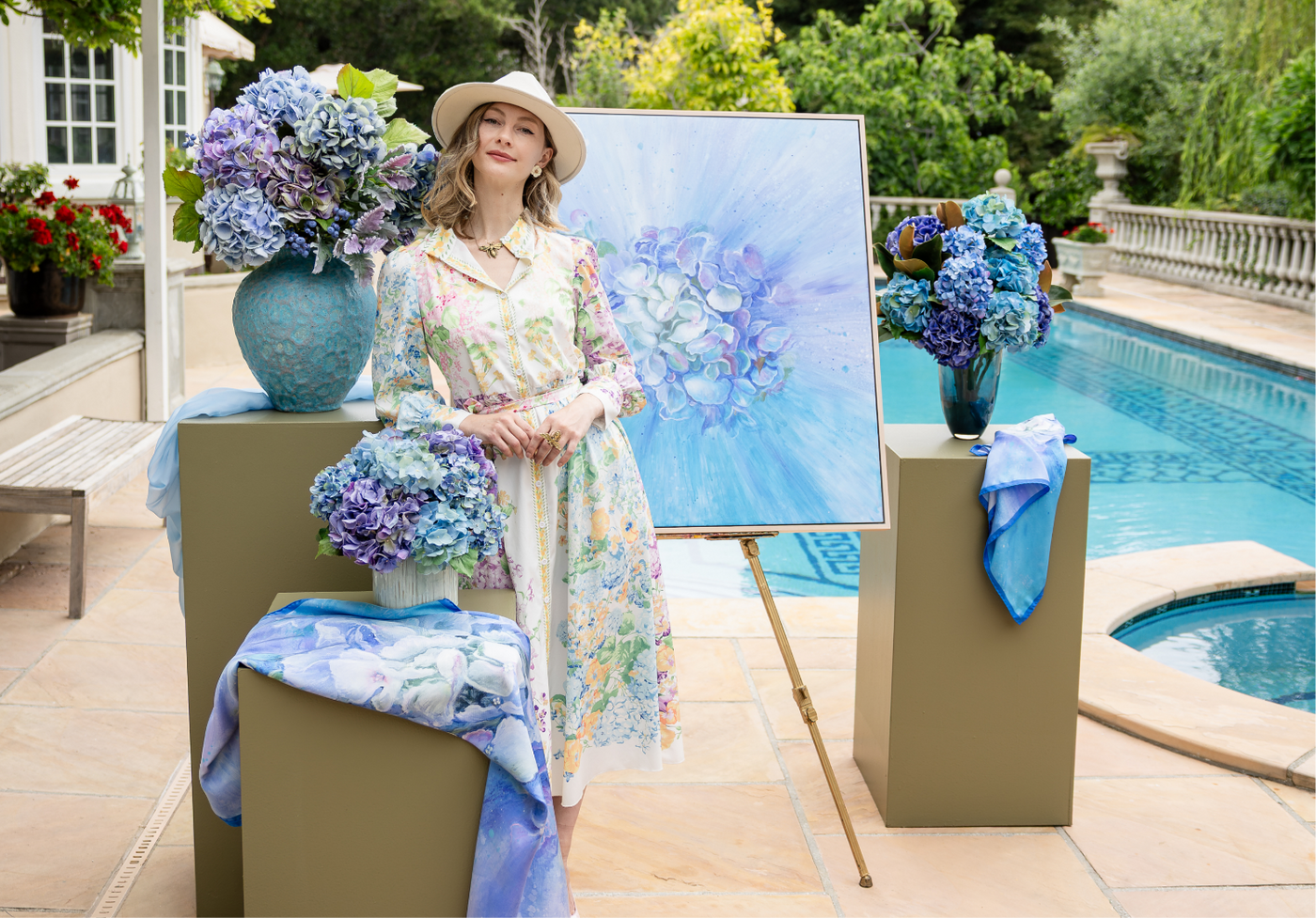 Woman in a floral dress standing next to a Hydrangea wall art, three hydrangea florals, and scarves by a pool.